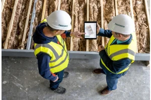 Twee vakmannen met helm en veiligheidshesje bekijken een bouwplan op een tablet naast glaswol isolatie tussen houten regels